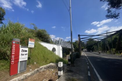 Street view with a metal pedestrian bridge, power pole, and lush green bushes under a blue sky with scattered clouds, on the right a roadway with scooters.
