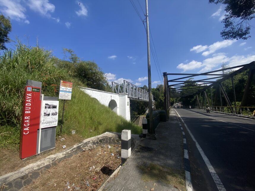 Street view with a metal pedestrian bridge, power pole, and lush green bushes under a blue sky with scattered clouds, on the right a roadway with scooters.