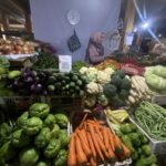Night market vegetable stall with colorful produce like carrots, peppers, cauliflower, cabbage, and a vendor wearing a hijab in the background.