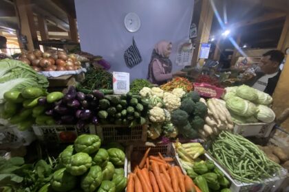 Night market vegetable stall with colorful produce like carrots, peppers, cauliflower, cabbage, and a vendor wearing a hijab in the background.