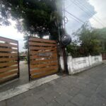 Wooden slat gate with a round sign by a white wall, set along a tree-lined street with overhead power lines.