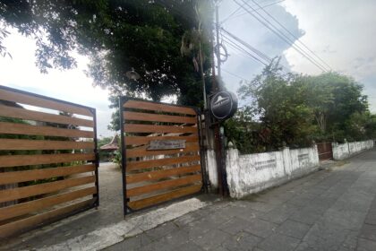 Wooden slat gate with a round sign by a white wall, set along a tree-lined street with overhead power lines.