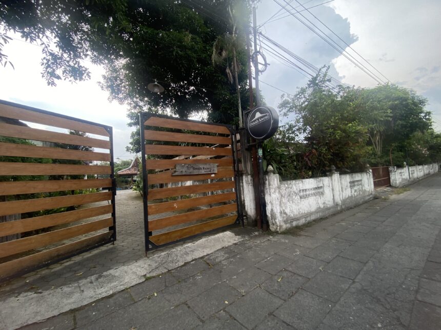 Wooden slat gate with a round sign by a white wall, set along a tree-lined street with overhead power lines.