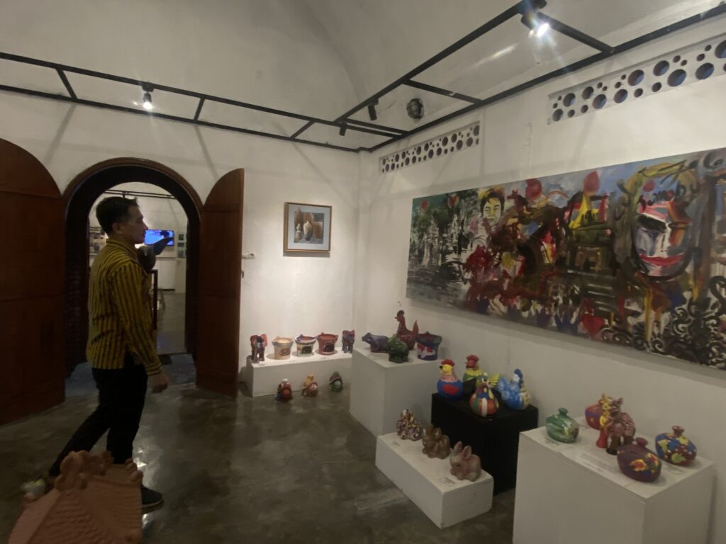 IMG_8216 - Mabur.co Museum gallery with a large colorful mural, framed art, and white display plinths holding ceramic pots; a man in a striped shirt walks through left side.