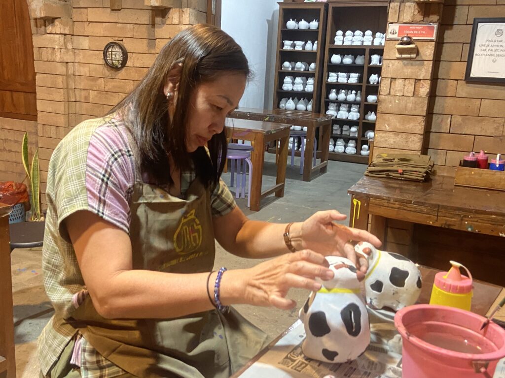 IMG_8234 - Mabur.co Woman painting a white skull with black spots at a wooden table in a rustic workshop.