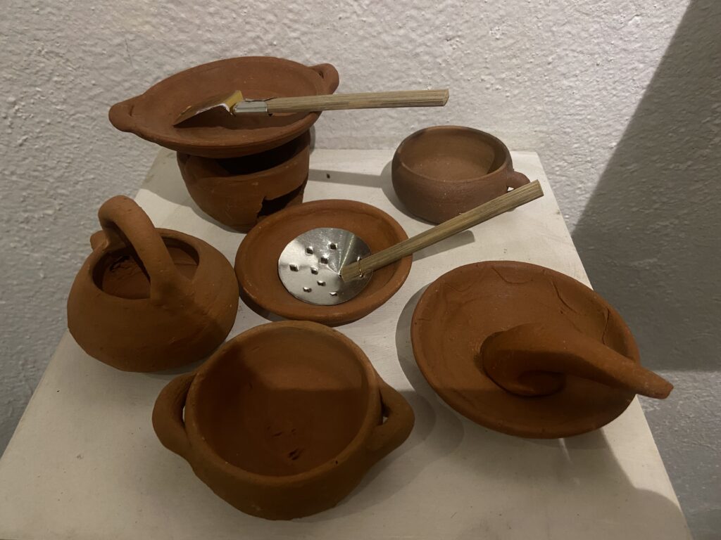 IMG_8252 - Mabur.co Group of small unglazed terracotta pots and lids arranged on a white pedestal, with wooden sticks resting across them as part of an sculpture.