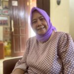 Smiling elderly woman wearing a purple headscarf and a patterned maroon top, indoors in a warmly lit room with framed pictures on the wall behind her.