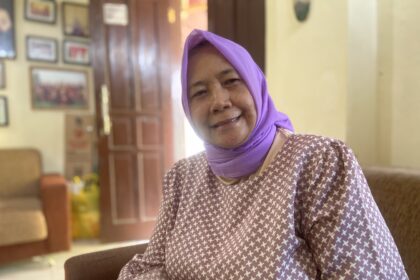 Smiling elderly woman wearing a purple headscarf and a patterned maroon top, indoors in a warmly lit room with framed pictures on the wall behind her.