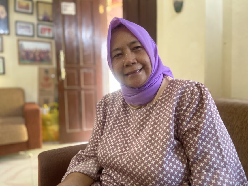 Smiling elderly woman wearing a purple headscarf and a patterned maroon top, indoors in a warmly lit room with framed pictures on the wall behind her.