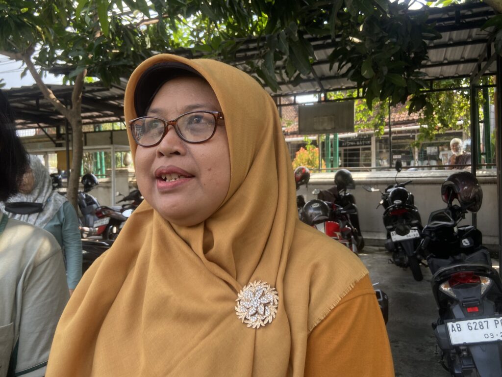 IMG_8344 - Mabur.co Close-up of a woman in a mustard hijab and glasses outdoors, with parked motorcycles and greenery in the background.