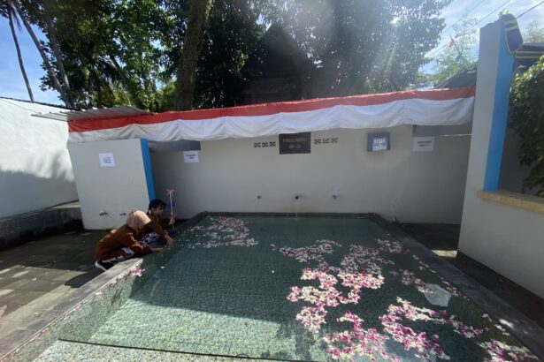 Two people kneeling by a shallow pool in a courtyard, petals scattered on a turquoise mosaic floor under a red-and-white canopy.