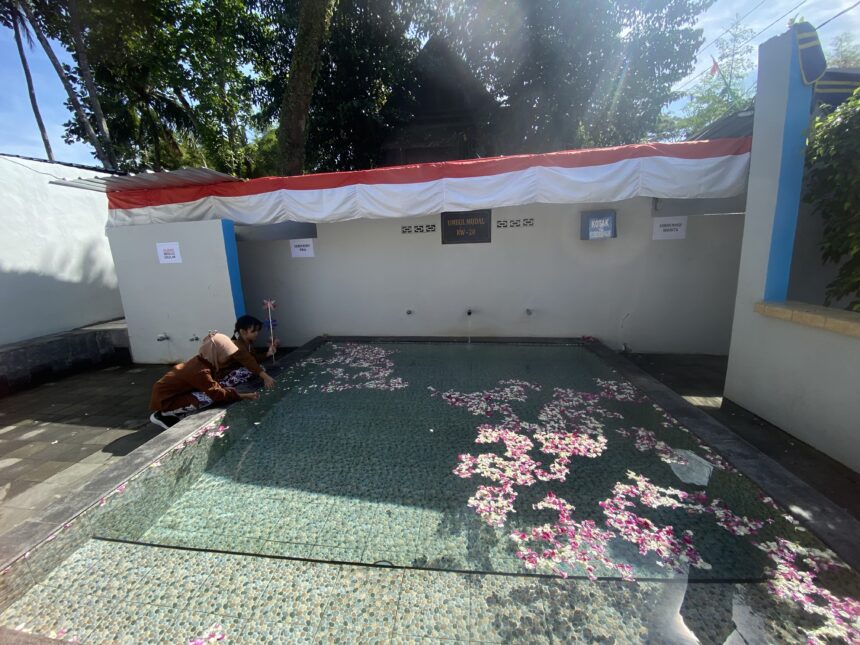 IMG_8430 - Mabur.co Two people kneeling by a shallow pool in a courtyard, petals scattered on a turquoise mosaic floor under a red-and-white canopy.