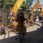 Dancers in colorful traditional costumes with masks and bell anklets perform in a street parade under yellow banners.