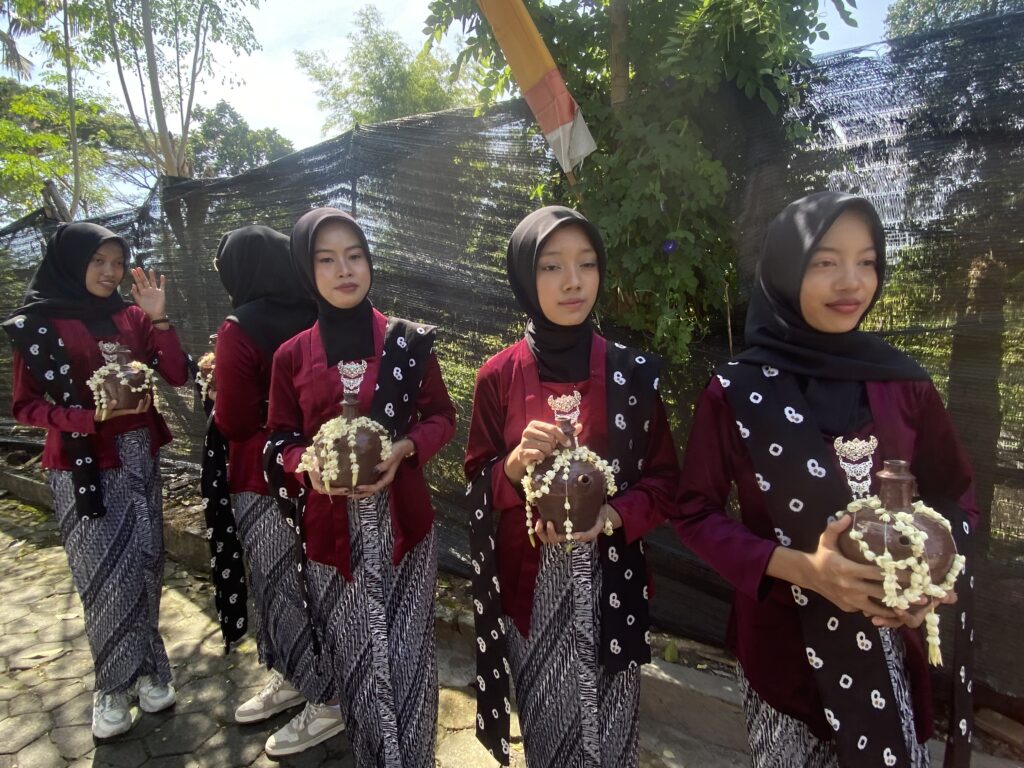 IMG_8448 1 - Mabur.co Group of women in matching maroon outfits and black hijabs stand outdoors, each holding a decorated pot with white flower garlands.