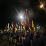 Nighttime parade with uniformed marchers carrying colorful tall flags in a row under streetlights, conveying a ceremonial procession.
