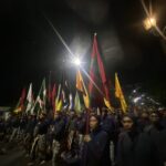 Row of uniformed marchers carrying colorful flags during a night parade along a street, with a large crowd watching.