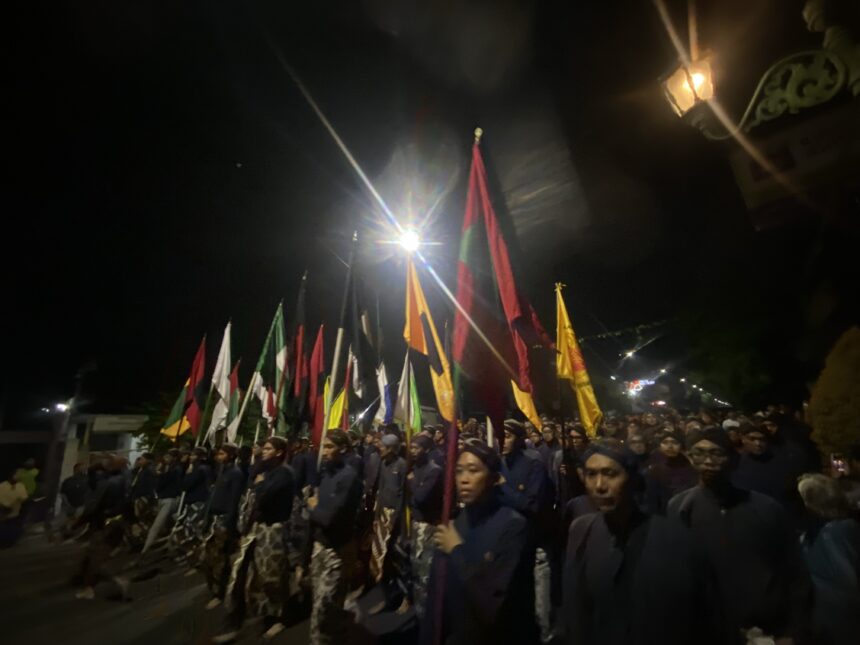 Row of uniformed marchers carrying colorful flags during a night parade along a street, with a large crowd watching.