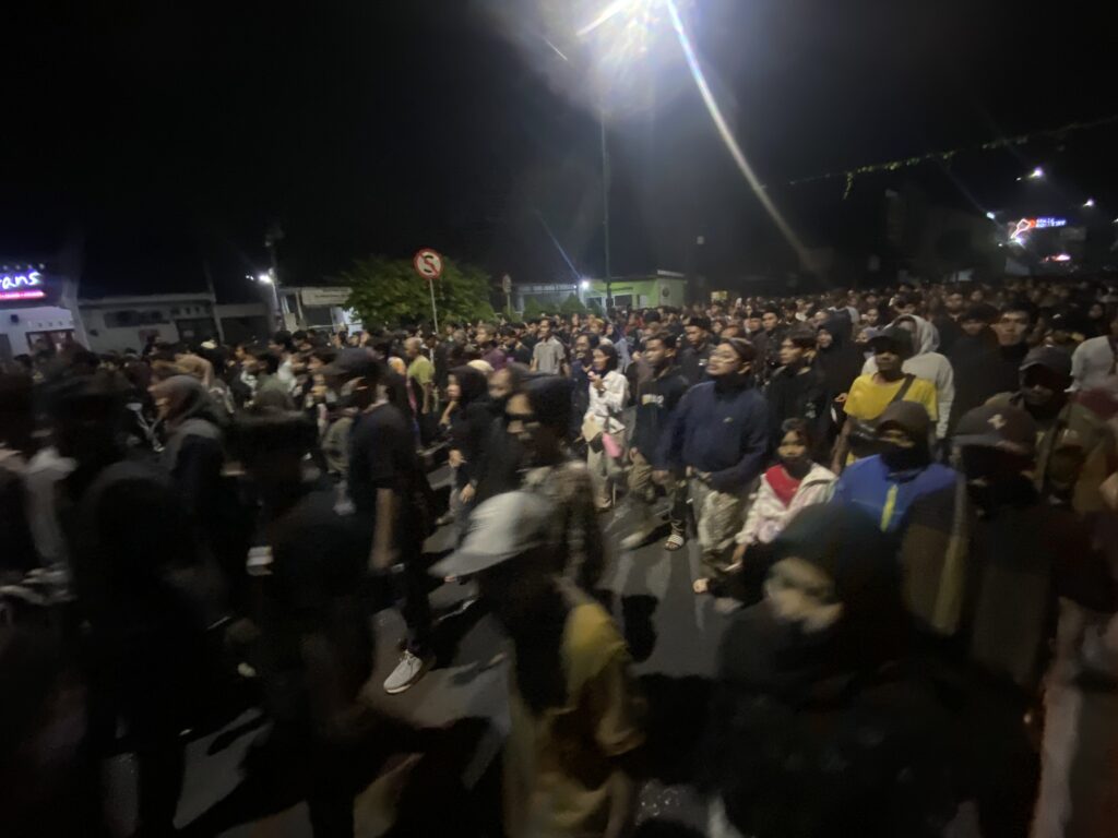 Large crowd of people walking down a city street at night under bright streetlights.