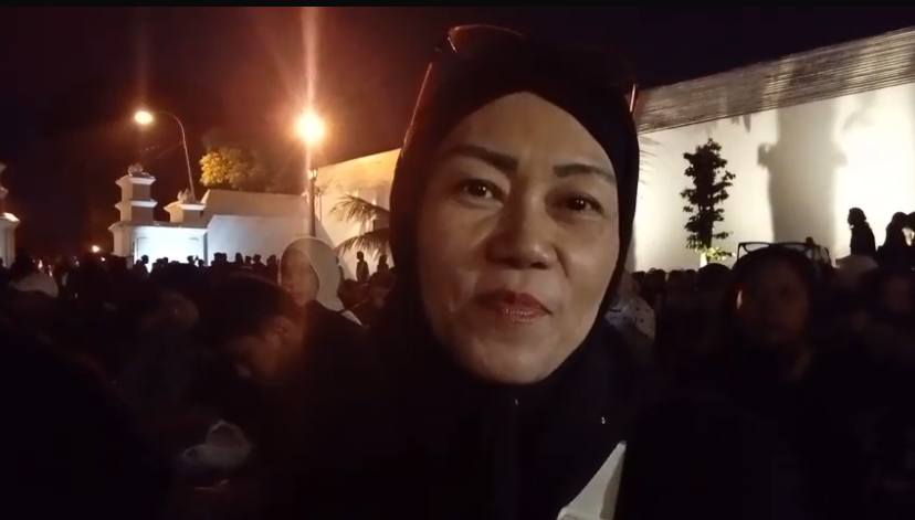 Close-up of a woman wearing a black hijab speaking to camera at night, surrounded by a large crowd near a lit wall.