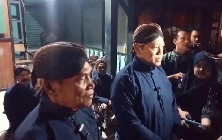 Two elderly men in dark blue traditional outfits and songkoks stand side by side at a press event, surrounded by reporters and onlookers indoors.