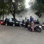 Parking lot with cars and many motorcycles beside a white wall, shaded by trees, and a person in a blue top and patterned skirt near the bikes.