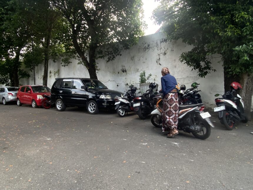 Parking lot with cars and many motorcycles beside a white wall, shaded by trees, and a person in a blue top and patterned skirt near the bikes.