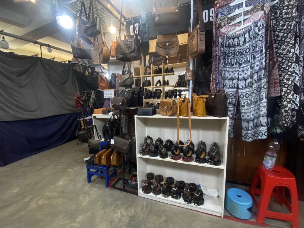 IMG_8578 - Mabur.co Indoor market stall displaying hanging leather bags and patterned garments; shelves hold belts, purses, and sandals behind a fabric backdrop.