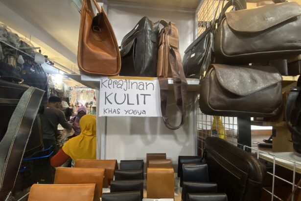 Shelf of leather bags in brown and black at a market stall, with a handwritten sign reading 'Kerajinan Kulit Khas Yogya' in the background.