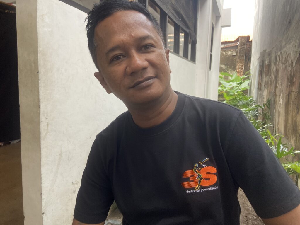 IMG_8589 - Mabur.co Portrait of a man in a black T-shirt with an orange graphic, standing in a narrow outdoor alley and looking at the camera, smiling subtly.