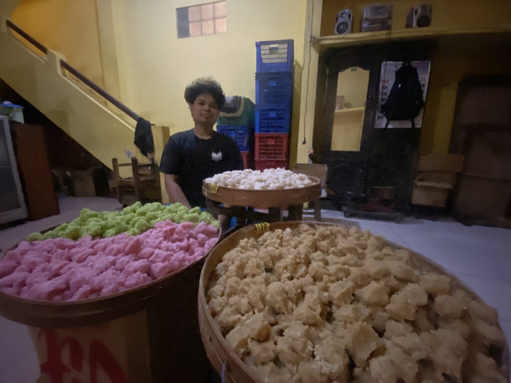 IMG_8601 - Mabur.co Person with curly hair standing behind two large baskets of pink and white puffed snacks in an indoor setting.