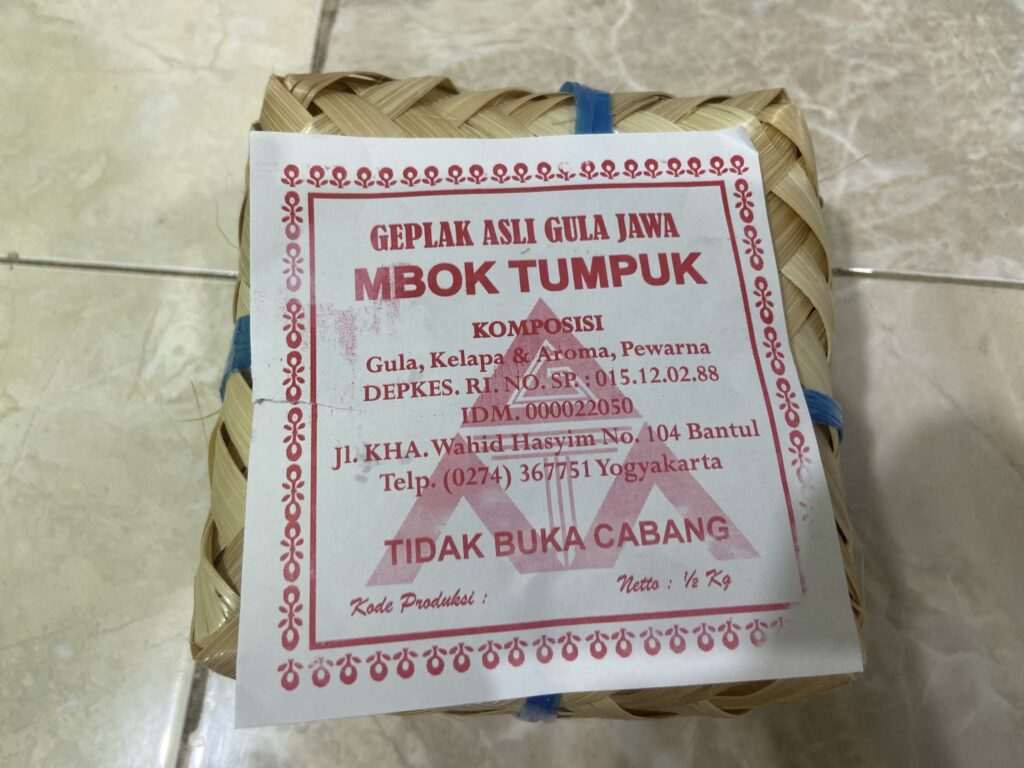 IMG_8609 - Mabur.co Woven palm-leaf package with a red and white label showing Indonesian text for a traditional snack (Gepak Asli Gula Jawa Mbok Tumpuk). It's taped and tied with blue plastic.