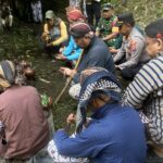 Group of Indonesian villagers and officials kneeling in a forested outdoor area, performing a traditional ceremony with brown clay pots.