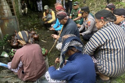 Group of Indonesian villagers and officials kneeling in a forested outdoor area, performing a traditional ceremony with brown clay pots.