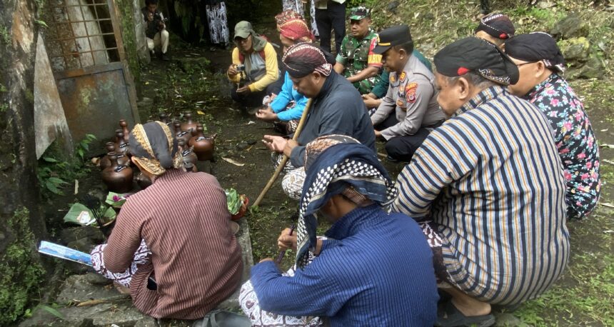 Group of Indonesian villagers and officials kneeling in a forested outdoor area, performing a traditional ceremony with brown clay pots.