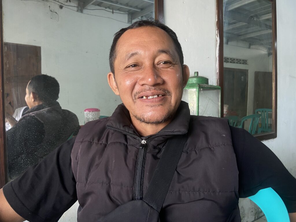 Smiling man in a black jacket posing in front of a glass window; reflection visible in the pane.