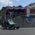 Green open-top jeep carrying people on a street near a patterned wall and official building with a banner and signage in Indonesian.