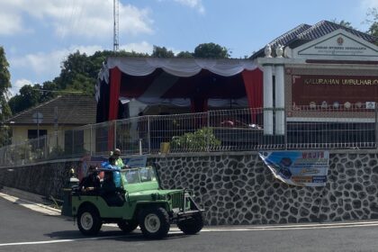Green open-top jeep carrying people on a street near a patterned wall and official building with a banner and signage in Indonesian.
