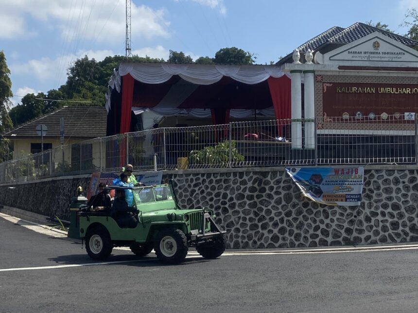 Green open-top jeep carrying people on a street near a patterned wall and official building with a banner and signage in Indonesian.