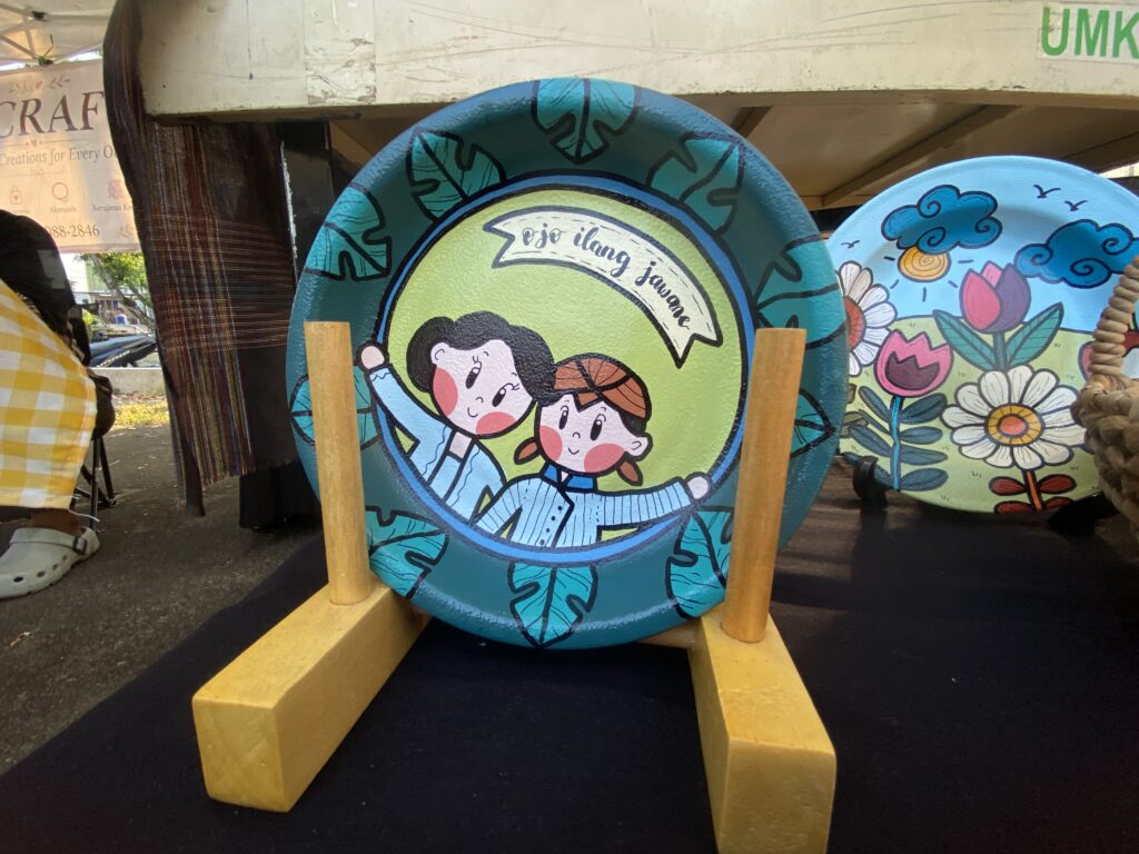 Round decorative panel with cartoon children in blue and green, mounted on a wooden stand at an outdoor market stall