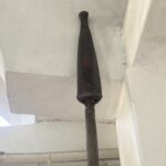 Vertical dark metal/wooden post rising from floor to ceiling in an indoor space, with white walls and exposed pipes around it.