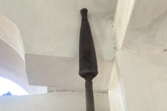 Vertical dark metal/wooden post rising from floor to ceiling in an indoor space, with white walls and exposed pipes around it.