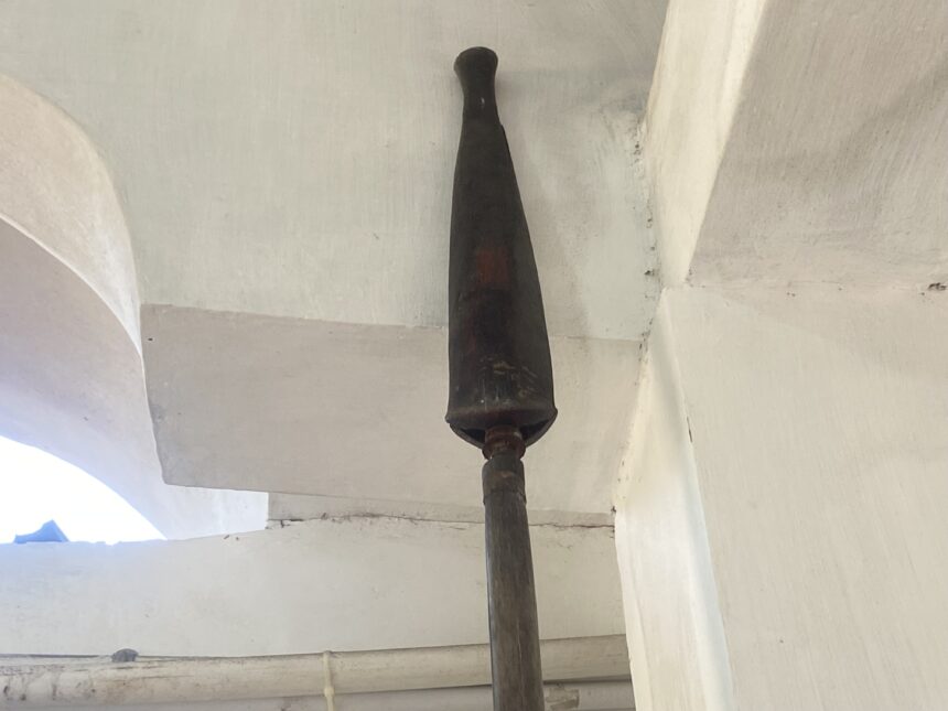 Vertical dark metal/wooden post rising from floor to ceiling in an indoor space, with white walls and exposed pipes around it.