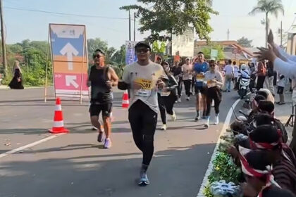 Runners participate in a road race while spectators cheer from the sidewalk on a sunny day.