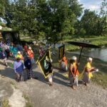 A colorful cultural procession with people in traditional attire walking along a roadside beside a canal, carrying banners and flags.