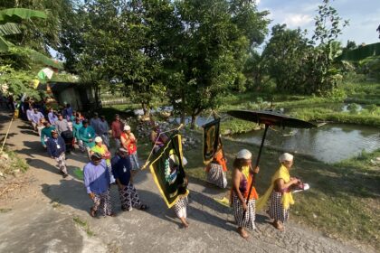 A colorful cultural procession with people in traditional attire walking along a roadside beside a canal, carrying banners and flags.