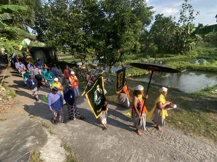 IMG_8751 - Mabur.co A colorful cultural procession with people in traditional attire walking along a roadside beside a canal, carrying banners and flags.