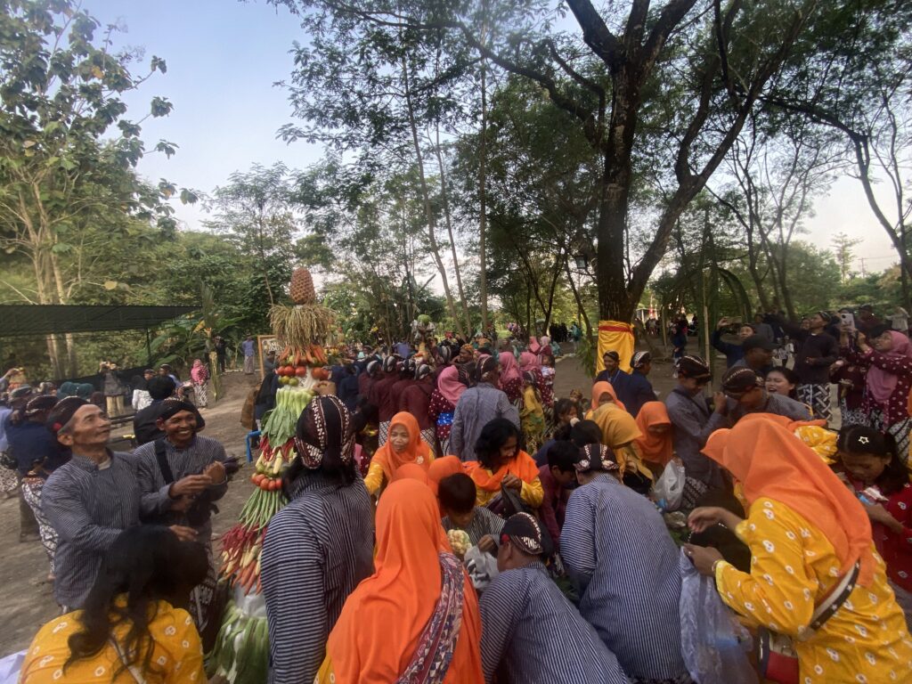 IMG_8775 - Mabur.co Crowd of people in blue-striped outfits and orange headscarves gather around a tall fruit-and-vegetable centerpiece at an outdoor festival, with trees in the background.