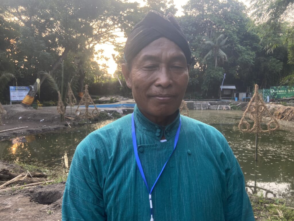 IMG_8786 - Mabur.co Close-up of a person wearing a teal traditional shirt and a black head wrap, standing by a pond with trees at sunset behind them.