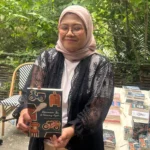Woman in a pink hijab and glasses proudly holding a book outdoors, with a table of books in the background.