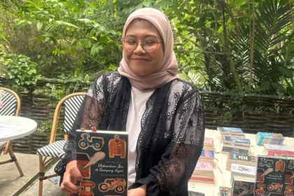 Woman in a pink hijab and glasses proudly holding a book outdoors, with a table of books in the background.